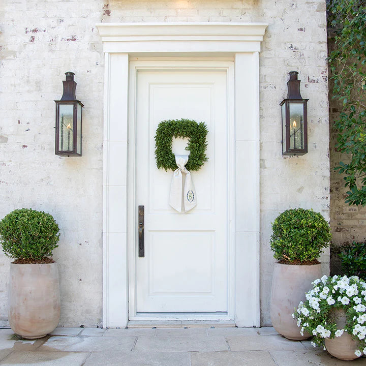 White door with decorative wreath, flanked by potted plants and wall sconces on a stone building.
