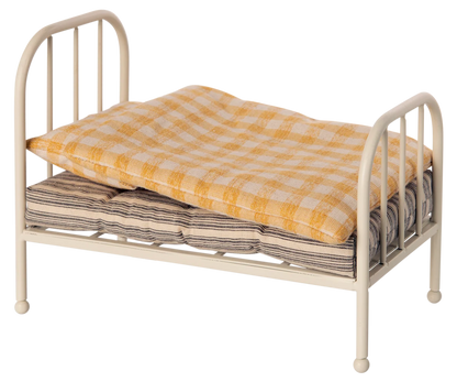 A small metal doll bed with a cream-colored frame, shown with a yellow-and-white checkered quilt and a striped mattress on a plain white background.