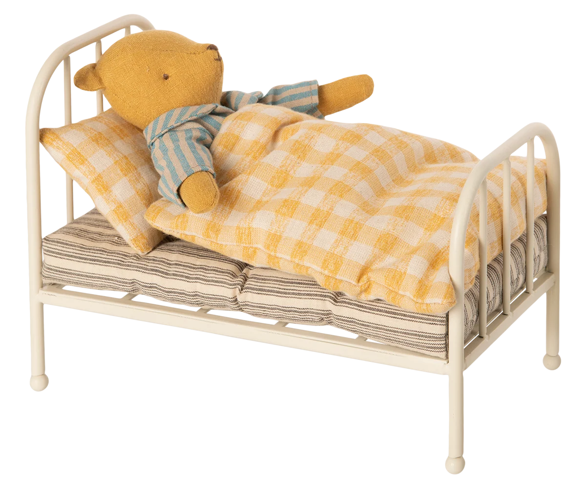 A small yellow teddy bear wearing blue and cream striped pajamas lies on a miniature cream-colored metal bed with a yellow-and-white checkered blanket and pillow, isolated on a white background.