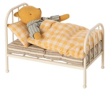 A small yellow teddy bear wearing blue and cream striped pajamas lies on a miniature cream-colored metal bed with a yellow-and-white checkered blanket and pillow, isolated on a white background.