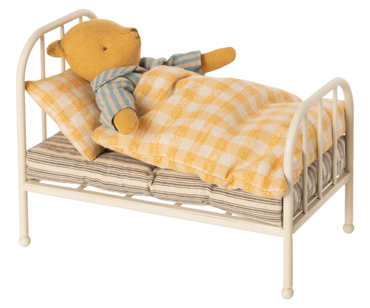 A small yellow teddy bear wearing blue and cream striped pajamas lies on a miniature cream-colored metal bed with a yellow-and-white checkered blanket and pillow, isolated on a white background.