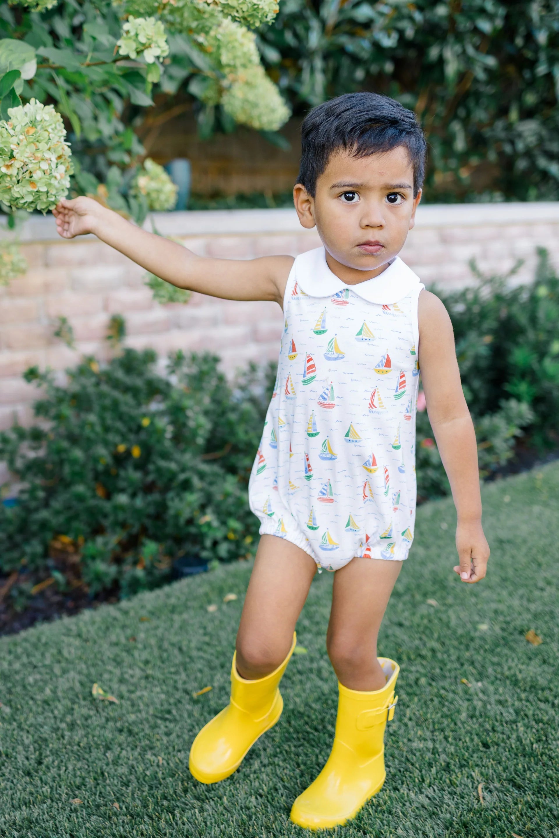 Child wearing a sleeveless dress with sailboat pattern and yellow rain boots outdoors.