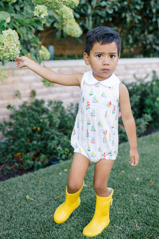 Child wearing a sleeveless dress with sailboat pattern and yellow rain boots outdoors.