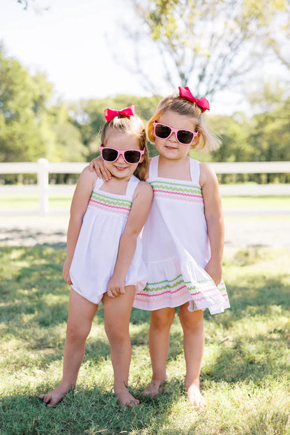 A white Sundress with multicolored ric rac trim and a bow at the back neck, designed for children.