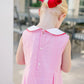 Child wearing a pink checkered dress with a red bow in the hair, standing outdoors.