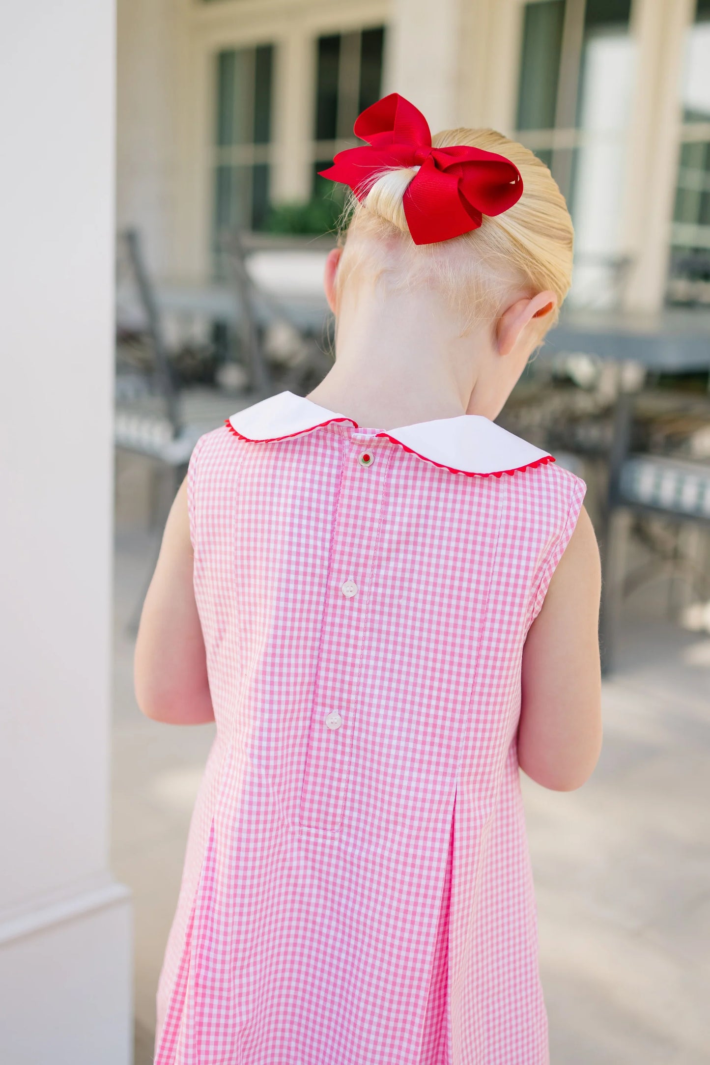Child wearing a pink checkered dress with a red bow in the hair, standing outdoors.